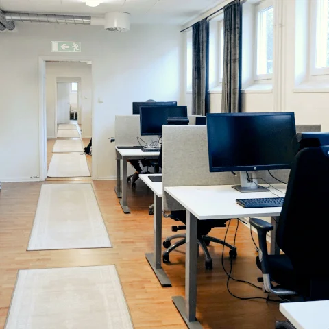 Ljust kontor med höj- och sänkbara skrivbord, datorskärmar och stolar placerade längs en korridor med trägolv