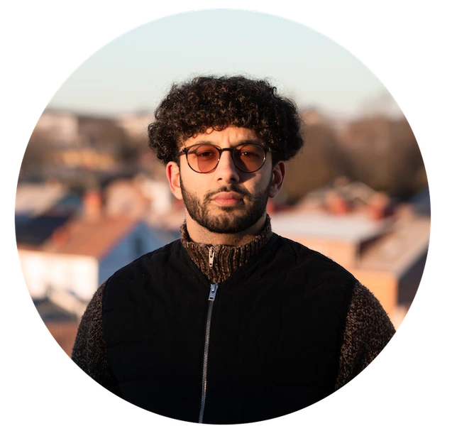 Porträtt av grundaren Robin Rushdi med glasögon och mörk väst, stående utomhus med suddiga takåsar i bakgrunden.