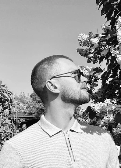 Svartvitt profilporträtt av William Janssen med solglasögon och ljus tröja med dragkedja vid halsen, stående utomhus bland växtlighet.